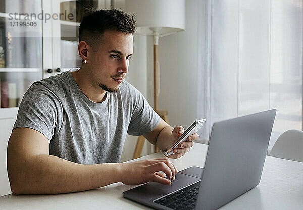Gut aussehender männlicher Unternehmer mit Smartphone und Laptop auf dem Tisch im Heimbüro