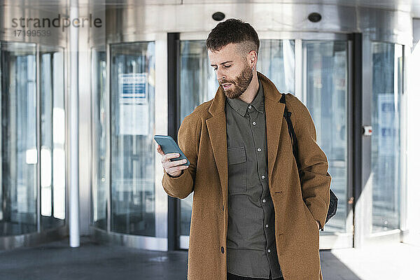 Geschäftsmann mit Jacke  der ein Mobiltelefon benutzt  während er mit den Händen in den Taschen vor einem Gebäude steht