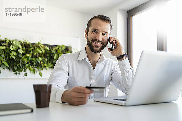 Lächelnder Geschäftsmann mit Kreditkarte und Laptop im Gespräch mit dem Handy im Büro