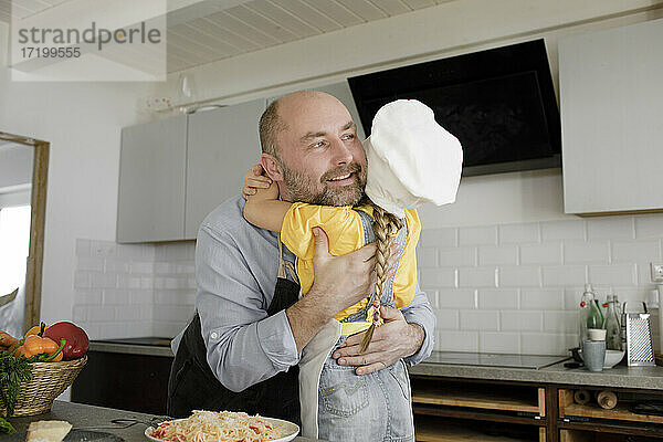 Vater umarmt seine Tochter  die eine Kochmütze trägt  während sie zu Hause in der Küche steht
