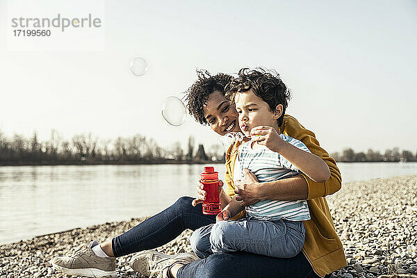Junge spielt mit einem Seifenblasenstab auf dem Schoß seiner Mutter an einem sonnigen Tag