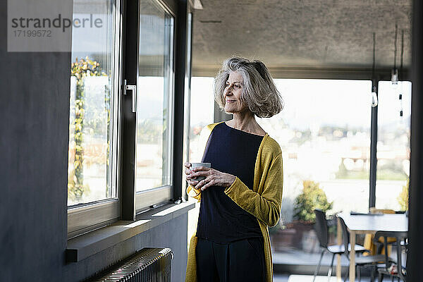 Lächelnde Frau mit Kaffeetasse  die durch das Fenster schaut  während sie zu Hause steht