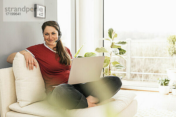 Lächelnde Frau mit Laptop und Kopfhörern  die auf dem Sofa im Wohnzimmer sitzt