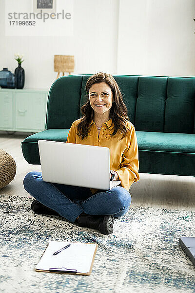 Lächelnde Frau mit Laptop auf dem Boden sitzend auf dem Sofa im Wohnzimmer