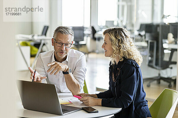Älterer Geschäftsmann mit Laptop  der einem Kollegen einen Geschäftsplan erläutert  während er am Tisch im Büro sitzt