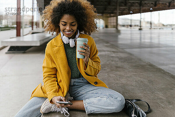 Lächelnde Afro-Frau mit Einweg-Kaffeebecher und Mobiltelefon in einer Sitzecke