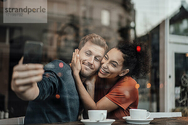 Freund nimmt Selfie mit Freundin durch Handy beim Sitzen im Cafe