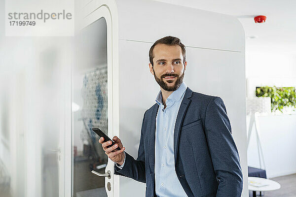 Geschäftsmann mit Mobiltelefon  der lächelnd an einer Telefonzelle im Büro steht