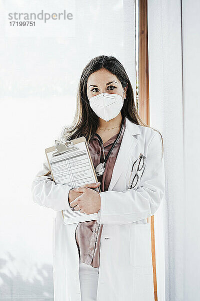 Weibliche medizinische Fachkraft mit Gesichtsschutzmaske  die ein Klemmbrett hält  während sie vor einem Glasfenster steht