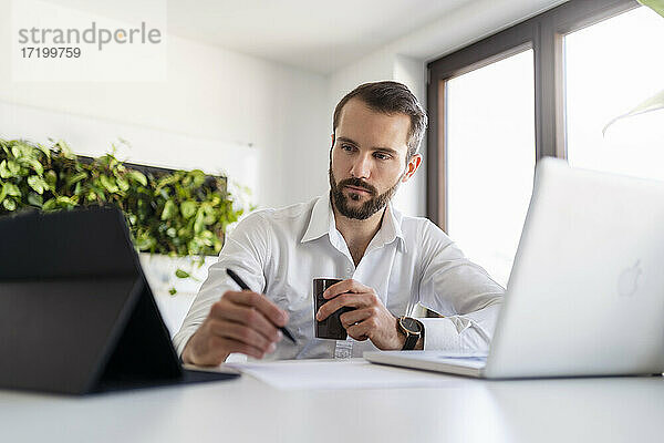 Geschäftsmann mit Laptop und Kaffeetasse bei der Nutzung eines digitalen Tablets im Büro