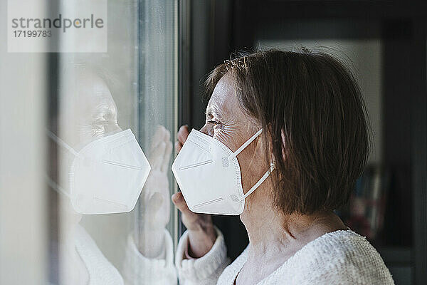 Ältere Frau mit Schutzmaske schaut während der Pandemie durch ein Fenster