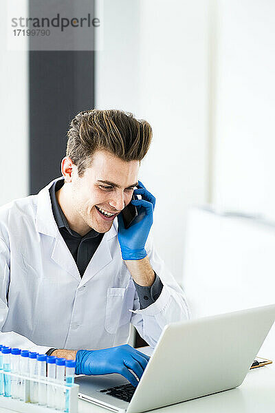 Lächelnder Wissenschaftler  der mit seinem Smartphone spricht  während er einen Laptop im Labor benutzt