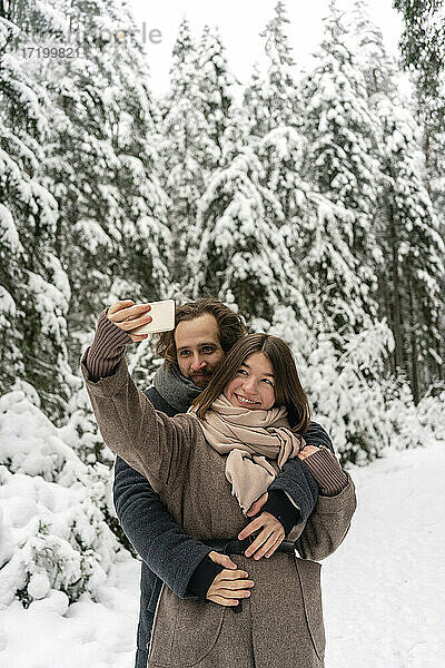 Freundin nimmt Selfie durch Handy mit Mann umarmt sie von hinten beim Stehen im Wald