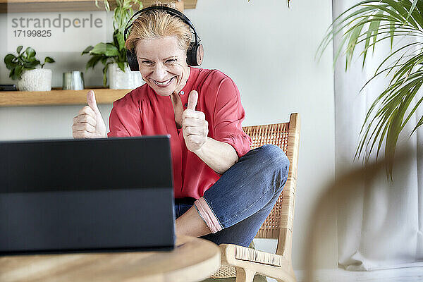 Glückliche reife Frau zeigt Daumen hoch Geste während Videoanruf auf Laptop zu Hause