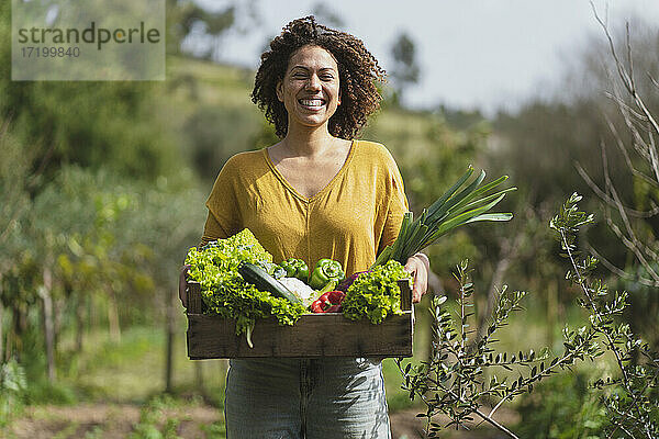 Lächelnde Frau  die im Garten stehend eine Kiste mit frischem Gemüse hält