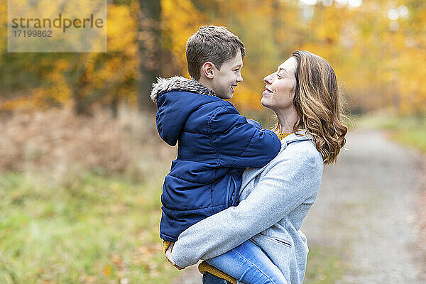 Mutter trägt lächelnden Sohn in den Armen  während sie im Wald steht