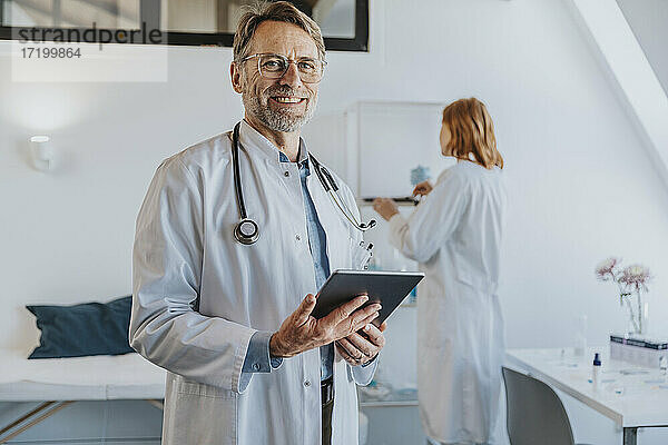 Lächelnder Arzt mit digitalem Tablet  der mit einem Mitarbeiter im Hintergrund in einer Klinik steht