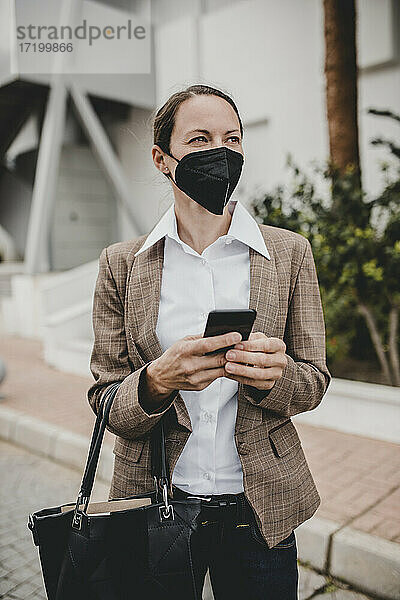 Geschäftsfrau mit Schutzmaske  die ein Smartphone in der Hand hält und an einem Gebäude steht  während sie wegschaut