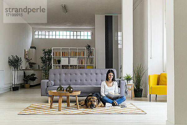 Lächelnde Frau liest ein Buch und sitzt mit ihrem Hund auf dem Teppich im Wohnzimmer