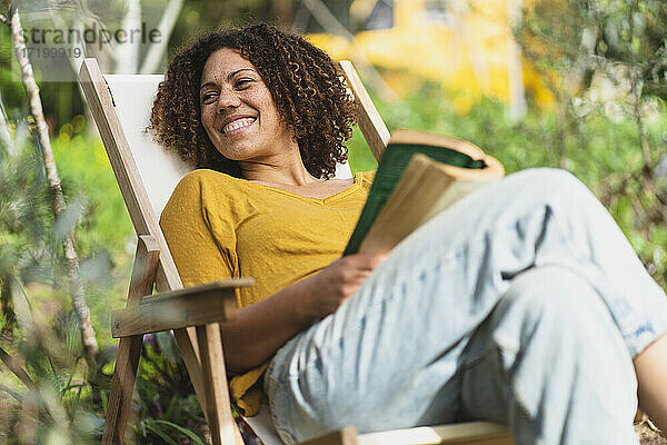 Lächelnde Frau mit lockigem Haar und Buch auf einem Stuhl im Garten sitzend