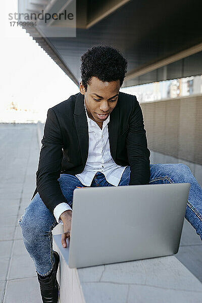 Männlicher Unternehmer  der einen Laptop benutzt  während er auf einer Stützmauer sitzt