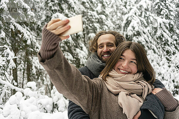 Lächelnde Frau  die ein Selfie mit einem Mann durch ein Mobiltelefon macht  während sie im Wald steht