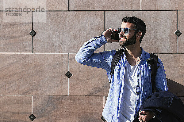 Junger Geschäftsmann  der an einem sonnigen Tag mit seinem Smartphone telefoniert  während er an der Wand steht