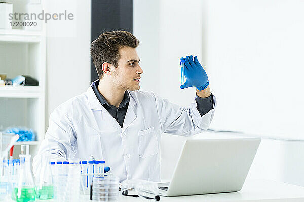 Männlicher Wissenschaftler  der eine Flüssigkeit im Reagenzglas betrachtet  während er im Labor an einem Laptop arbeitet