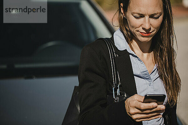 Lächelnde Geschäftsfrau mit Handtasche  die ihr Mobiltelefon an einem sonnigen Tag gegen ein Auto hält