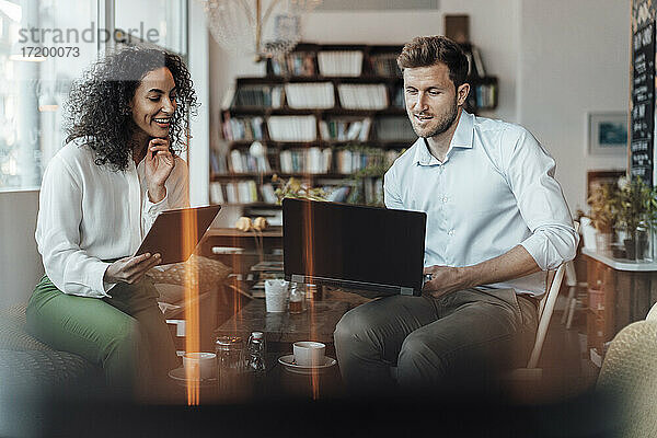 Lächelnde Geschäftsfrau mit digitalem Tablet  die neben einem Kollegen sitzt  der in einem Café am Laptop arbeitet