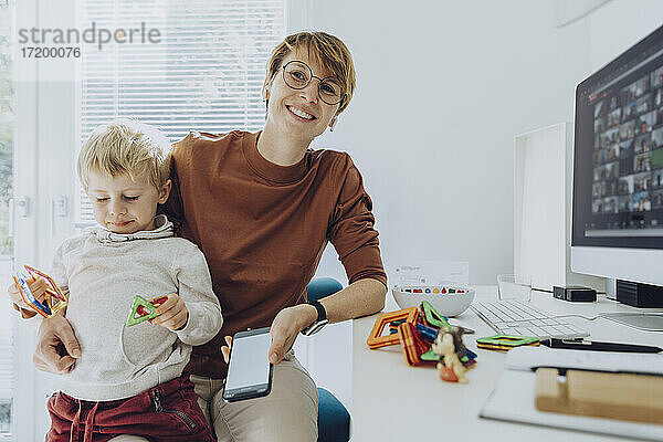 Lächelnde Mutter mit Sohn  der auf dem Schoß sitzt und mit Spielzeug im Büro zu Hause spielt
