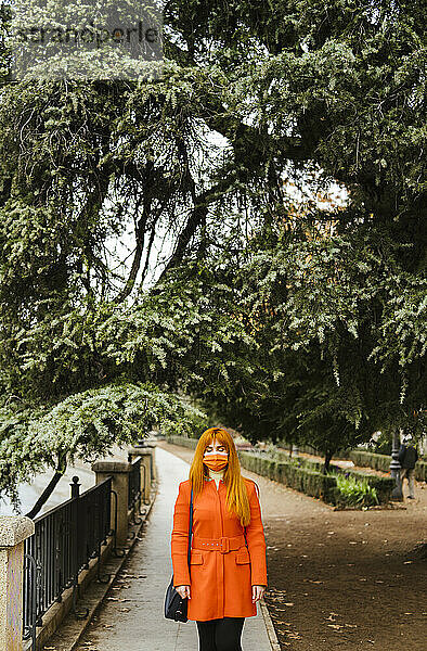 Frau mit Gesichtsschutzmaske  die in einem öffentlichen Park an einem Baum steht