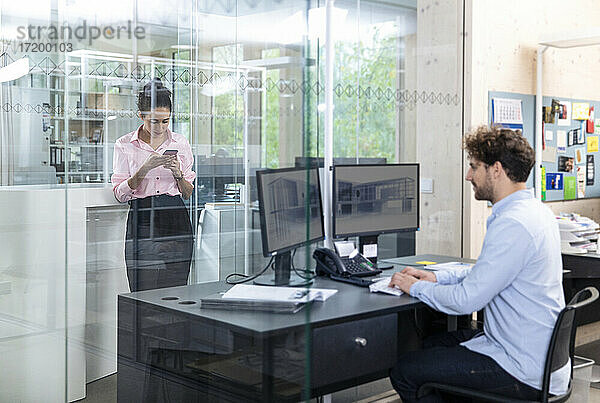 Junger männlicher Unternehmer bei der Arbeit  während eine Kollegin im Büro ein Smartphone benutzt