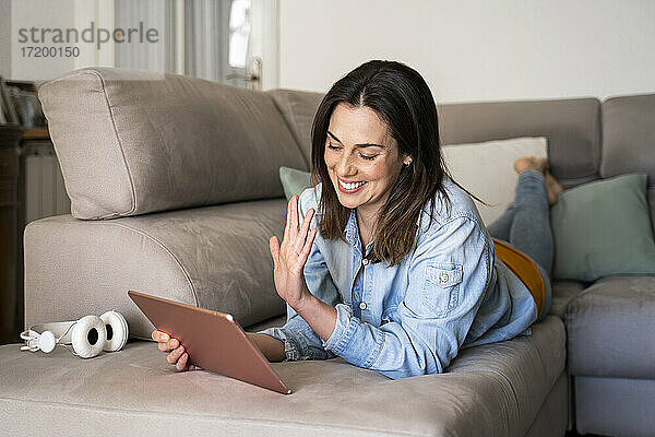 Glückliche Frau  die über ein digitales Tablet einen Videoanruf führt  während sie zu Hause auf dem Sofa liegt