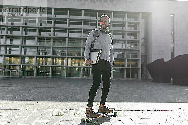 Männlicher Pendler mit Akten  der auf einem Skateboard vor einem Gebäude steht und wegschaut
