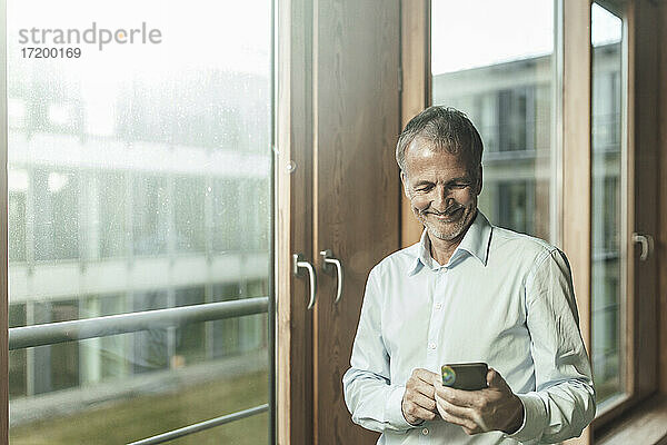 Lächelnder männlicher Unternehmer  der ein Smartphone benutzt  während er am Fenster im Büro steht