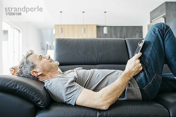Lächelnder reifer Mann mit digitalem Tablet  der auf dem Sofa im Wohnzimmer liegt und vor sich hin träumt