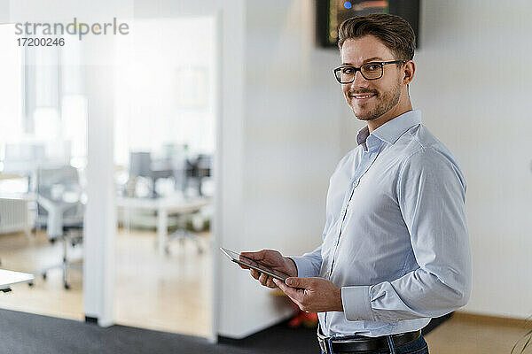 Lächelnder männlicher Unternehmer mit digitalem Tablet im Büro stehend