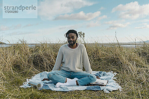 Junger Mann sitzt auf einer Decke inmitten von getrockneten Pflanzen gegen den Himmel