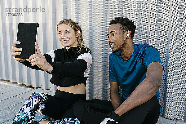 Junge Frau mit Mann nimmt Selfie durch digitale Tablette beim Sitzen auf dem Boden gegen die Wand
