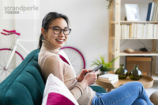 Junge Frau mit Brille sitzt mit Handy zu Hause