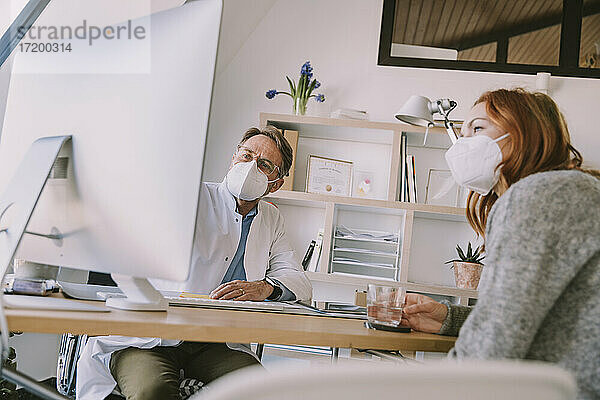 Arzt und Patient mit Gesichtsschutzmaske bei der Arbeit am Computer  während sie in einer Arztpraxis sitzen
