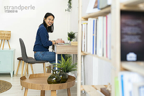 Frau lächelt  während sie an einem Laptop auf einem Tisch im Büro zu Hause sitzt