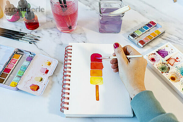 Frau malt Popsickle-Eis mit Aquarellfarbe in einem Buch auf dem Tisch