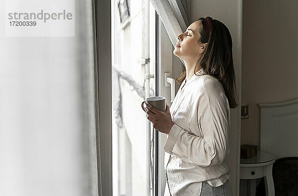 Entspannte junge Frau mit geschlossenen Augen  die eine Kaffeetasse hält  während sie am Fenster steht