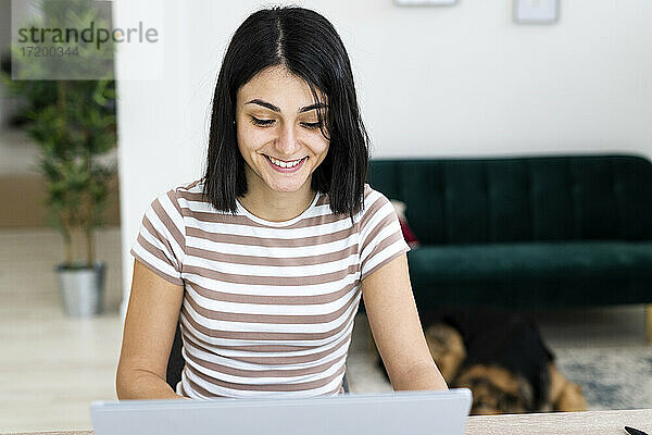 Lächelnde junge Frau  die einen Laptop benutzt  während sie am Tisch im Wohnzimmer sitzt