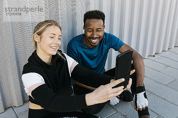 Fröhlicher männlicher und weiblicher Sportler  der ein Selfie durch ein digitales Tablet macht  während er auf dem Boden an der Wand sitzt