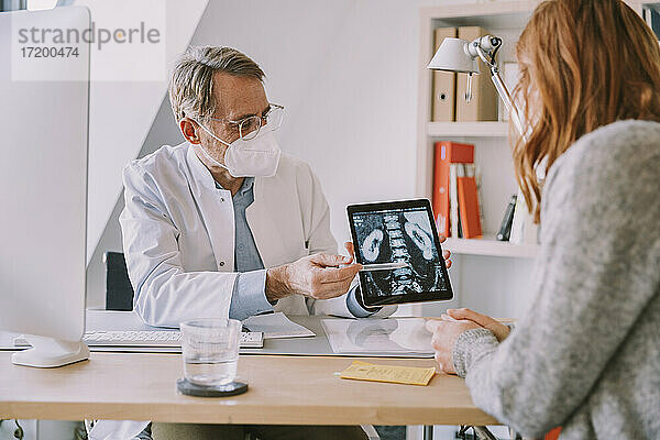 Arzt mit Gesichtsschutzmaske zeigt dem Patienten eine Röntgenaufnahme auf einem digitalen Tablet im Büro