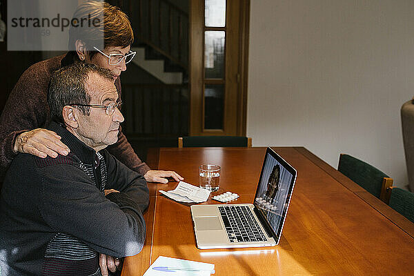 Eine Sozialarbeiterin steht neben einem älteren Mann  der sich mit einer Ärztin per Videoanruf über einen Laptop berät.
