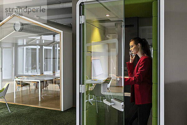 Lächelnde Unternehmerin  die einen Laptop benutzt  während sie in einer schalldichten Kabine im Büro mit einem Mobiltelefon spricht